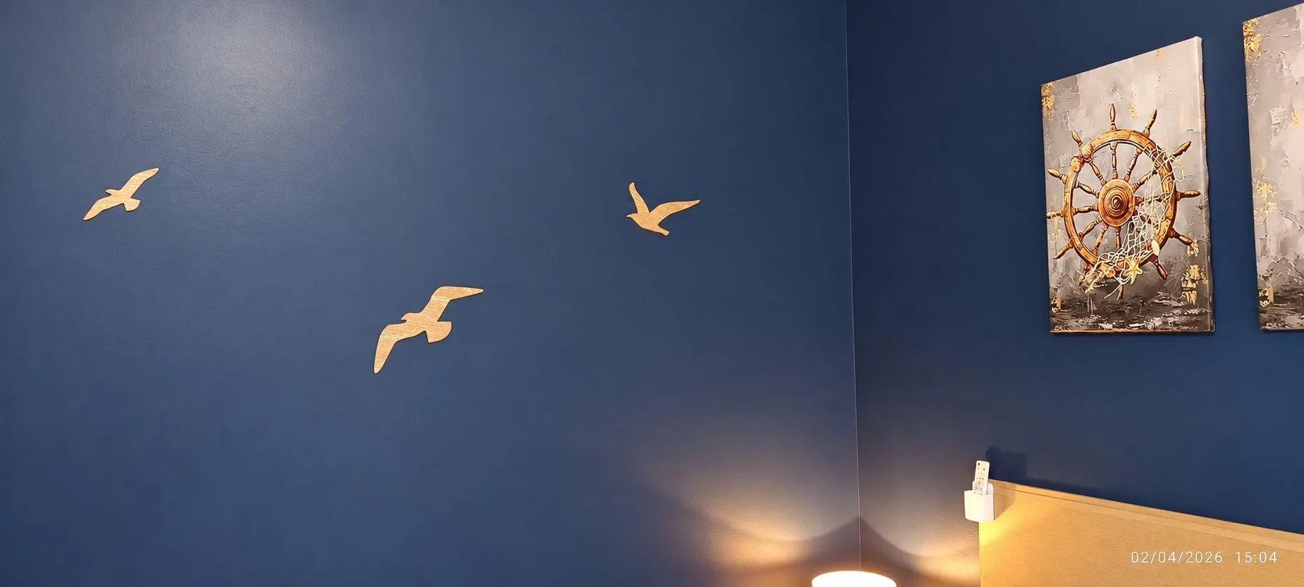 Un mur bleu marine décoré de trois mouettes dorées en vol et d'un tableau représentant une barre de bateau, avec une tête de lit jaune en bas à droite.