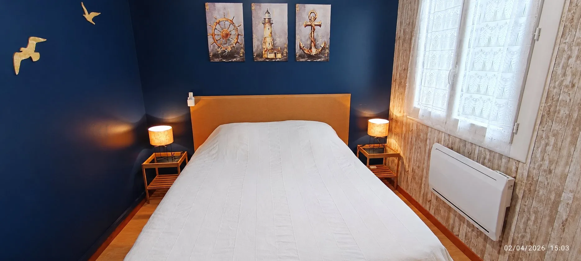 Chambre aux murs bleu marine avec un lit double, deux lampes de chevet allumées, trois tableaux marins et des mouettes décoratives.