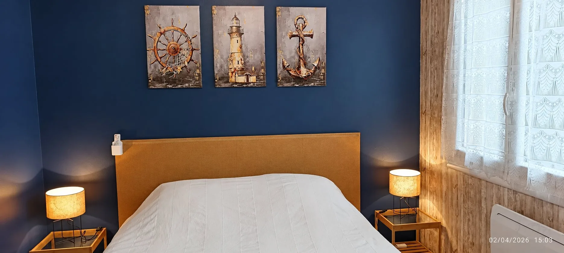 Une chambre aux murs bleu marine avec un lit double à tête de lit jaune, trois tableaux marins au-dessus, deux lampes de chevet allumées et une fenêtre aux rideaux en dentelle.