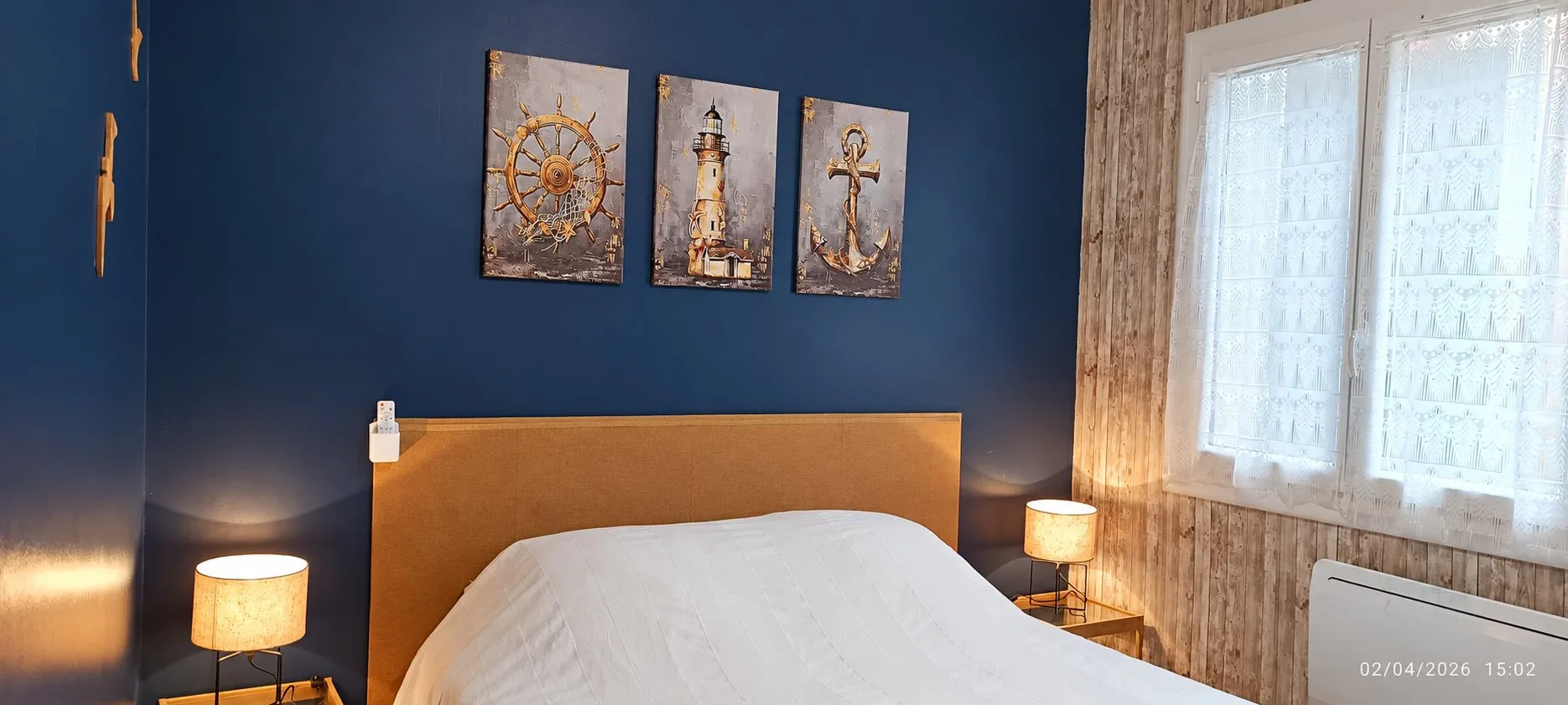 Chambre aux murs bleu marine avec trois tableaux marins au-dessus d'un lit double, flanqué de deux lampes allumées et d'une fenêtre à voilage blanc.