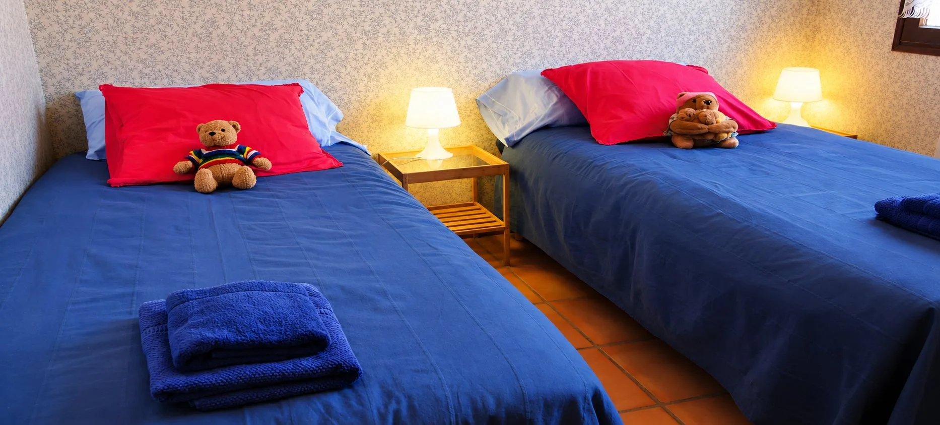 Une chambre avec deux lits couverts de draps bleus, des oreillers rouges ornés d'oursons en peluche, des serviettes pliées et une table de chevet en bois entre deux lampes allumées.