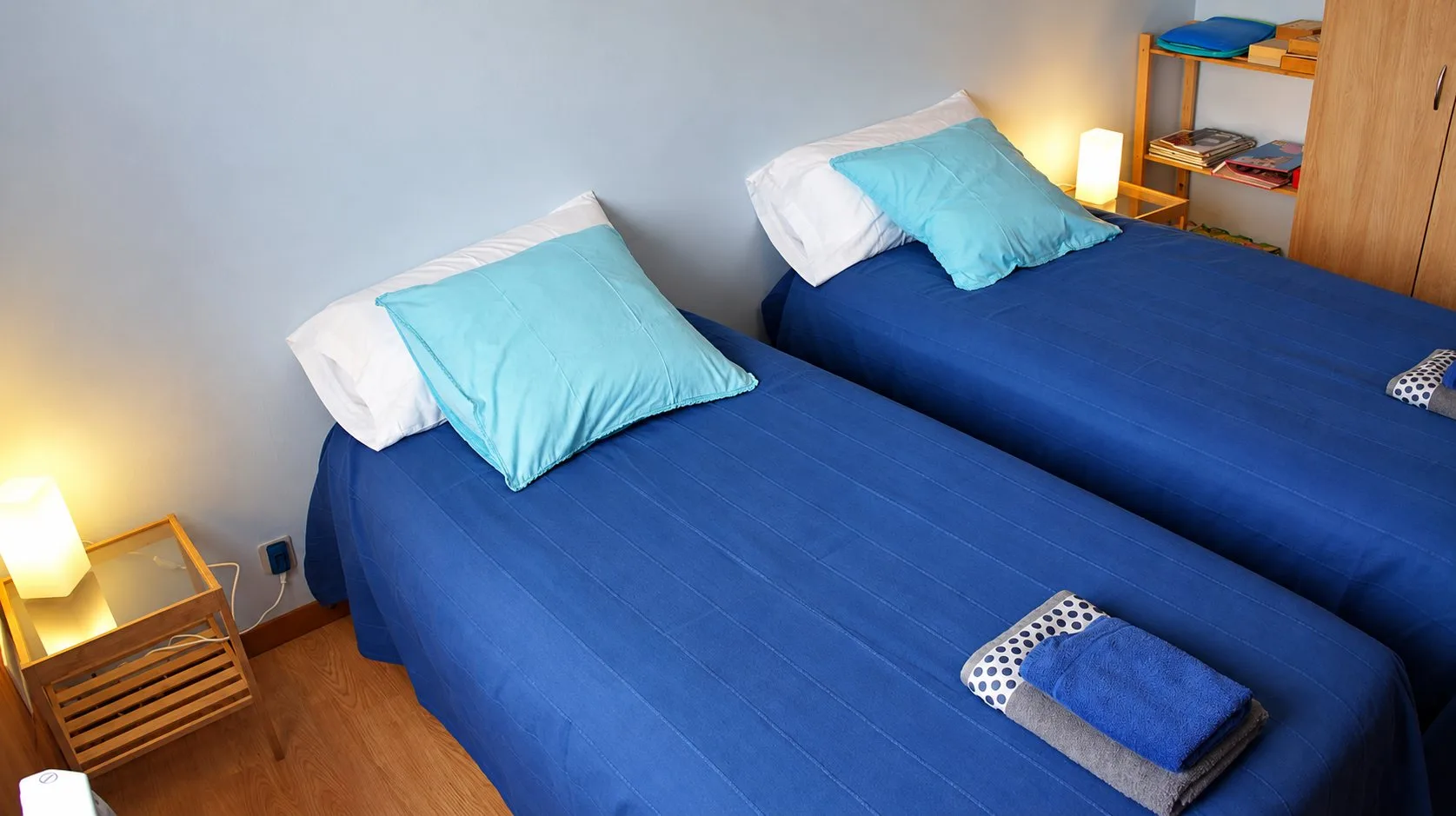 Chambre avec deux lits simples recouverts de couvre-lits bleus, coussins turquoise, lampes allumées et serviettes pliées sur un lit.