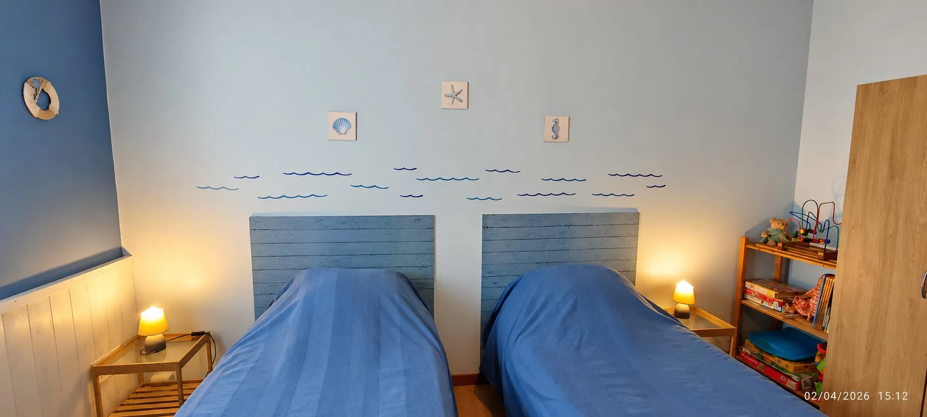 Chambre aux murs bleus avec deux lits simples aux têtes de lit en bois, des motifs de vagues et de coquillages au mur, deux lampes allumées et une étagère de jouets à droite.