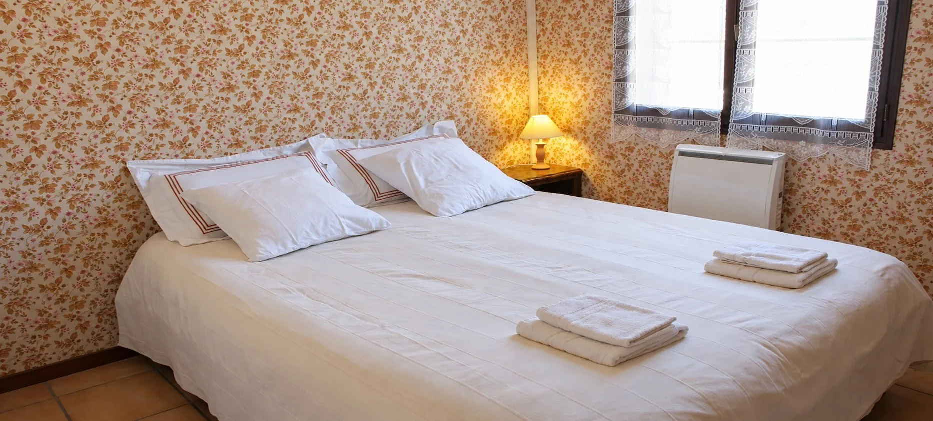 Une chambre avec un grand lit recouvert de blanc, des serviettes pliées, une lampe allumée et un papier peint à motifs floraux.