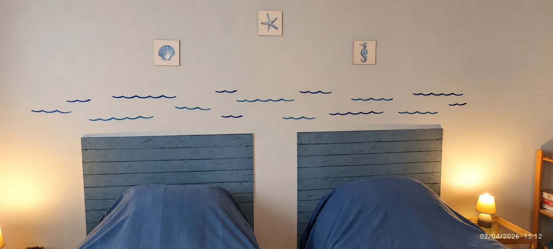 Chambre avec deux lits jumeaux à têtes de lit en bois bleu-gris, un mur décoré de petites vagues et de trois cadres représentant un coquillage, une étoile de mer et un hippocampe.