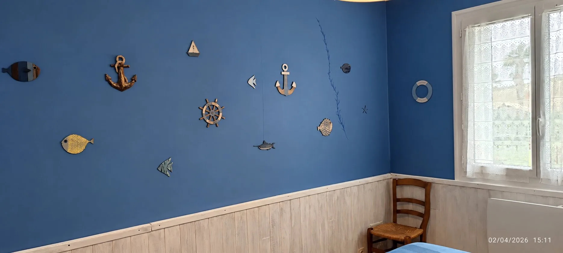 Chambre aux murs bleu marine ornés d'ancres, de poissons et d'une barre de bateau en bois, avec soubassement en lambris clair, une chaise en paille et une fenêtre aux rideaux blancs.