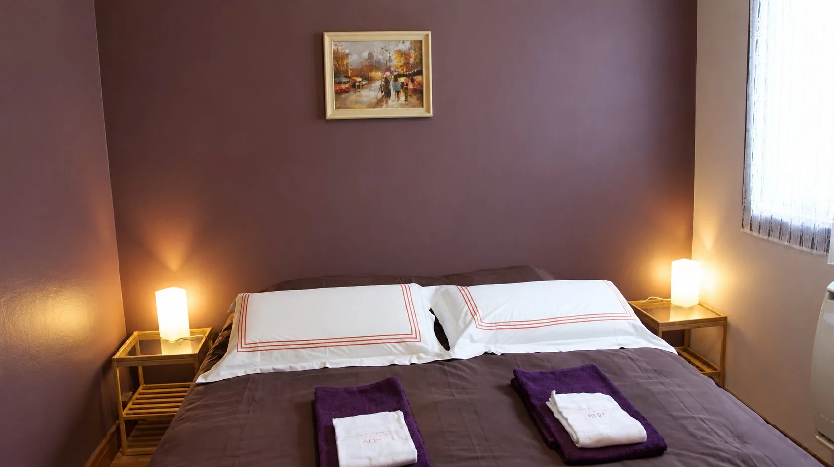 Chambre aux murs violet foncé avec un lit double, deux serviettes pliées sur le couvre-lit et deux lampes allumées sur des tables de chevet.