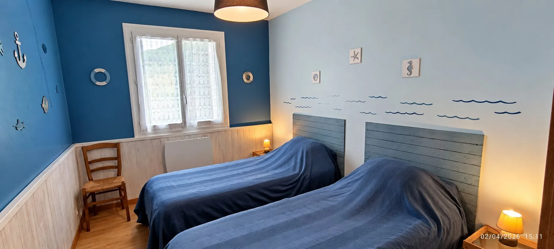 Chambre aux murs bleus décorés d'une ancre et de motifs marins, avec deux lits simples, des têtes de lit en bois bleu et des lampes de chevet allumées.