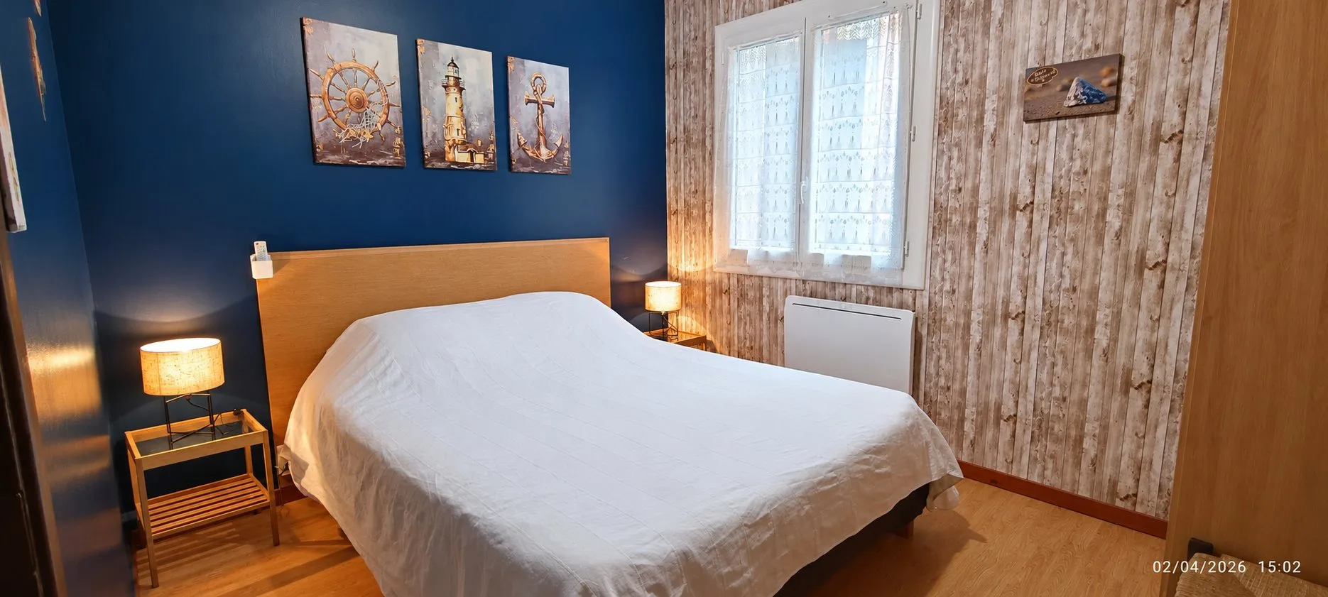 Chambre avec un grand lit double drapé de blanc, un mur bleu marine orné de trois tableaux marins, deux lampes de chevet et un mur en bois clair près de la fenêtre.