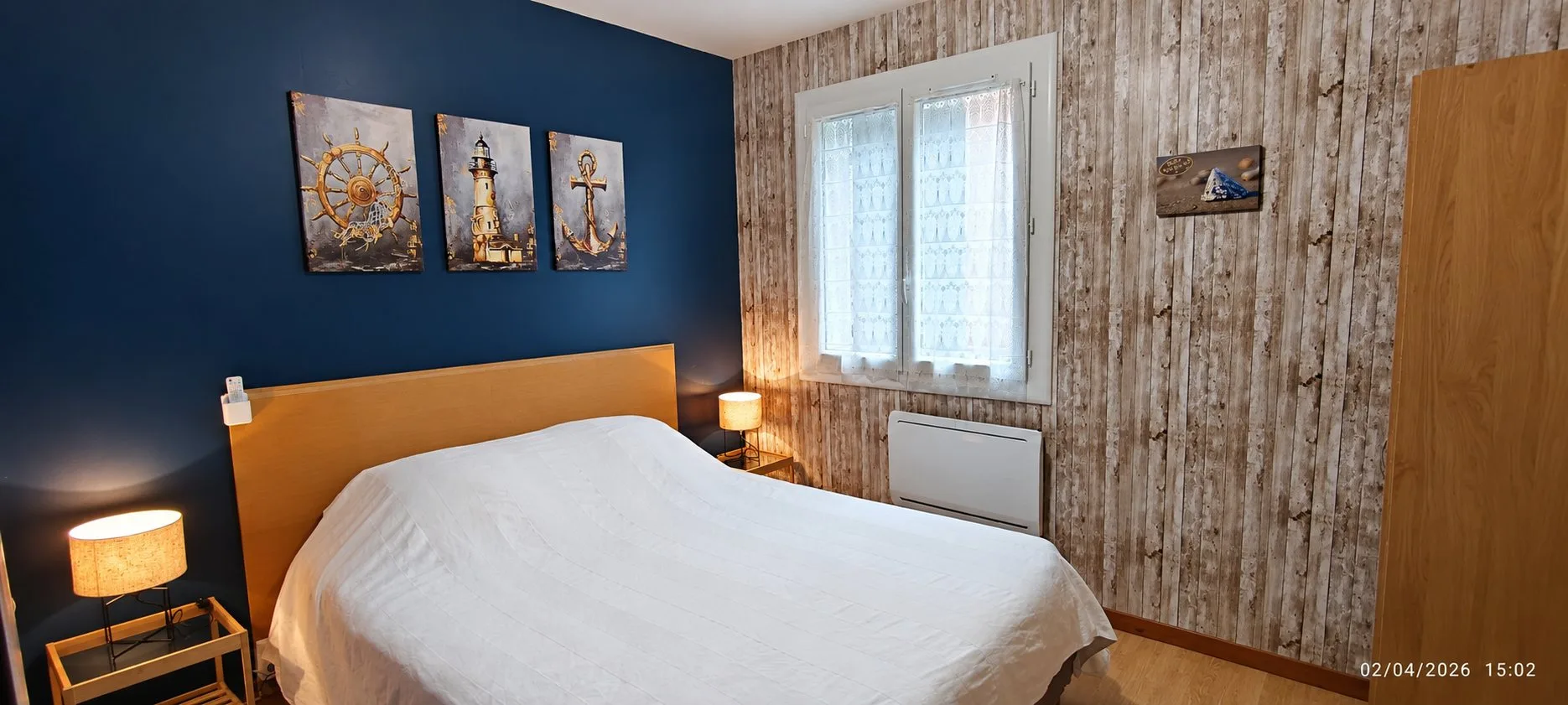 Chambre avec un grand lit double aux draps blancs, un mur bleu marine orné de trois tableaux marins, un mur imitation bois et deux lampes de chevet allumées.