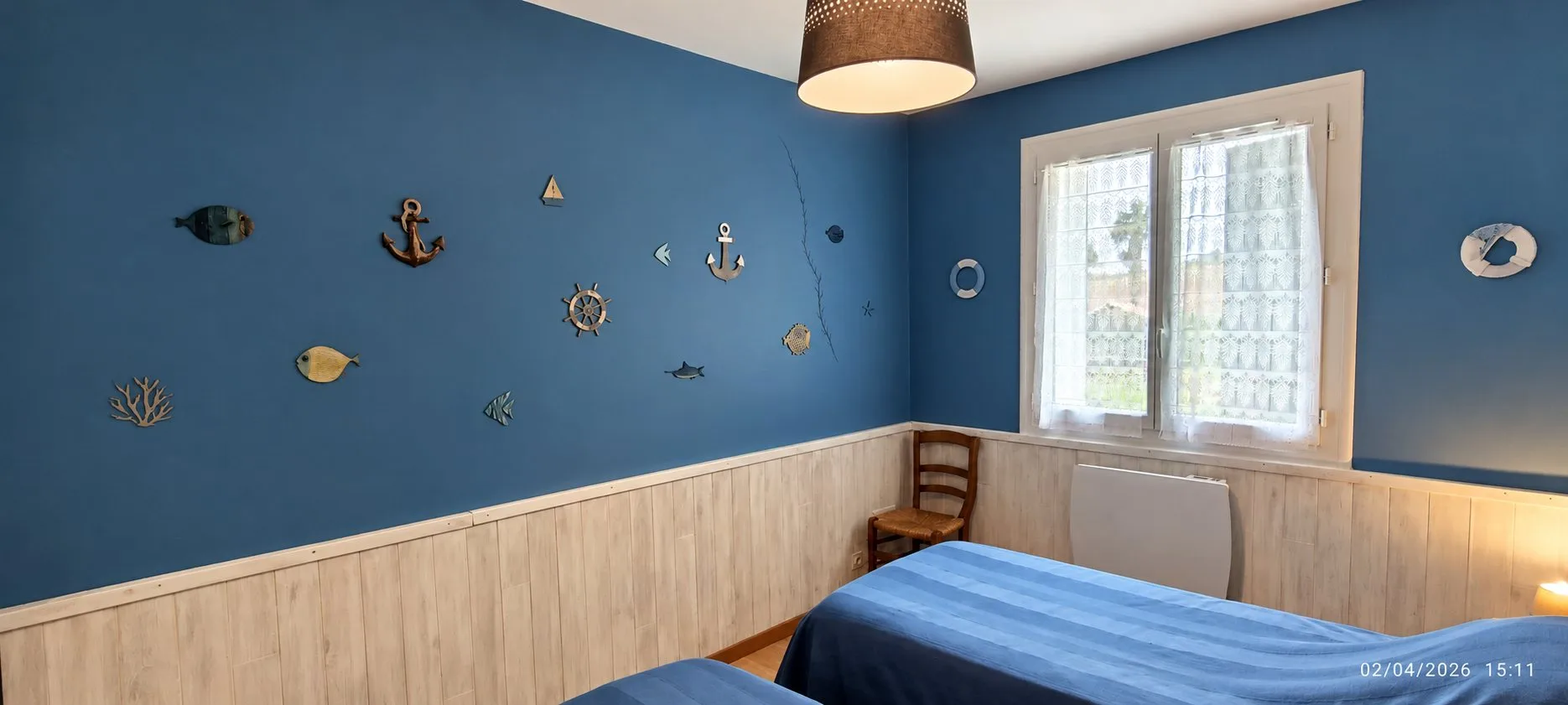 Chambre aux murs bleus décorés d'ancres, de poissons et d'une barre à roue, avec lambris clair, fenêtre à voilage et deux lits simples.