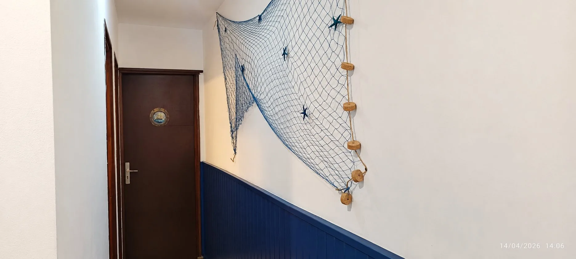 Un couloir aux murs clairs et à lambris bleu foncé, orné d'un filet de pêche bleu avec étoiles de mer et flotteurs en liège, menant à une porte en bois sombre.