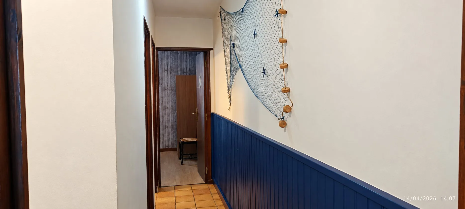 Un couloir aux murs clairs avec un lambris bleu marine, orné d'un filet de pêche avec flotteurs en liège et étoiles de mer, menant à une porte ouverte.