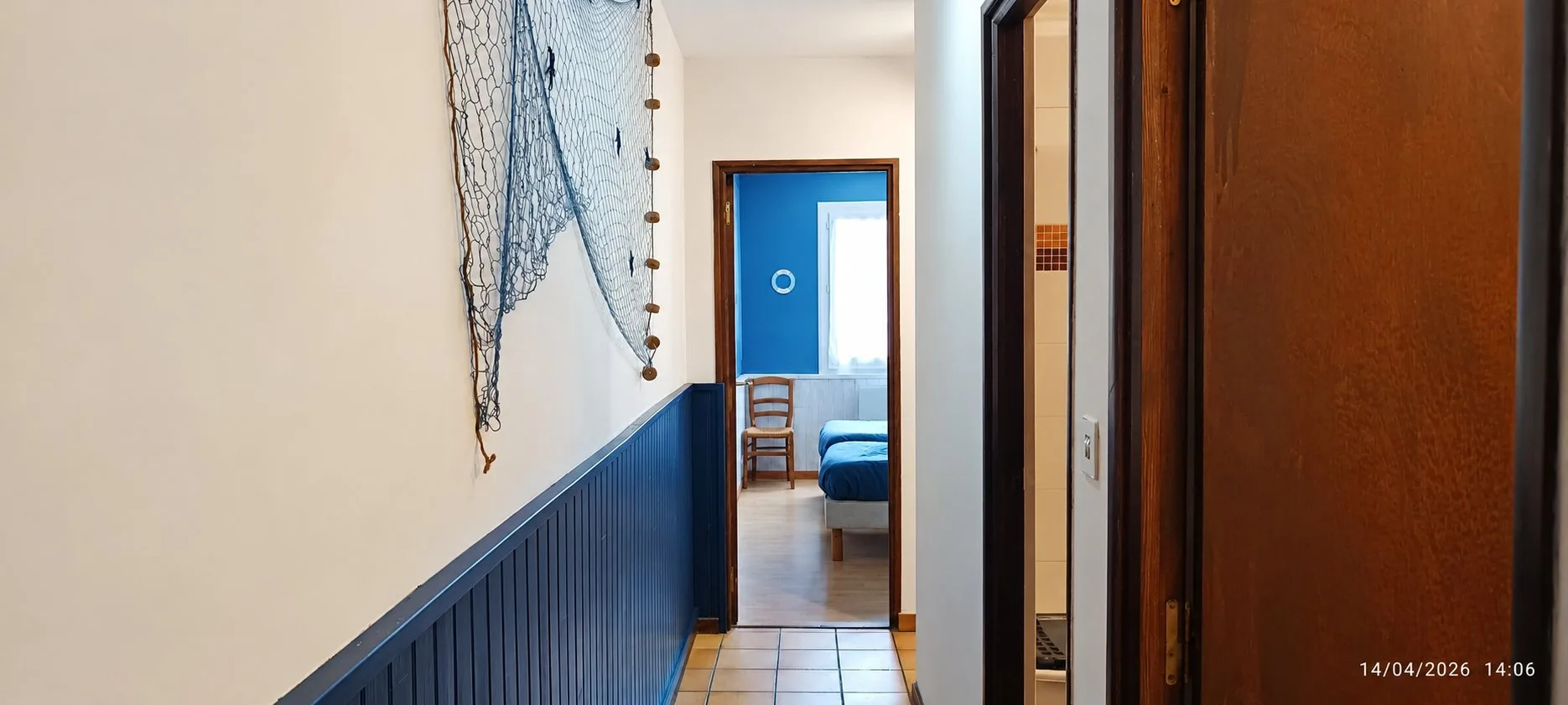 Un couloir aux murs crème et lambris bleu marine, décoré d'un filet de pêche, ouvre sur une chambre bleue avec lits et chaise en bois.