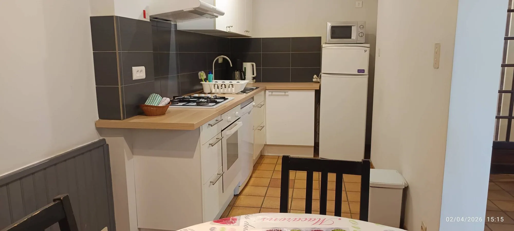 Petite cuisine équipée aux meubles blancs, avec plaque de cuisson à gaz, réfrigérateur, micro-ondes et carrelage mural gris foncé, à côté d'une table et d'une chaise.