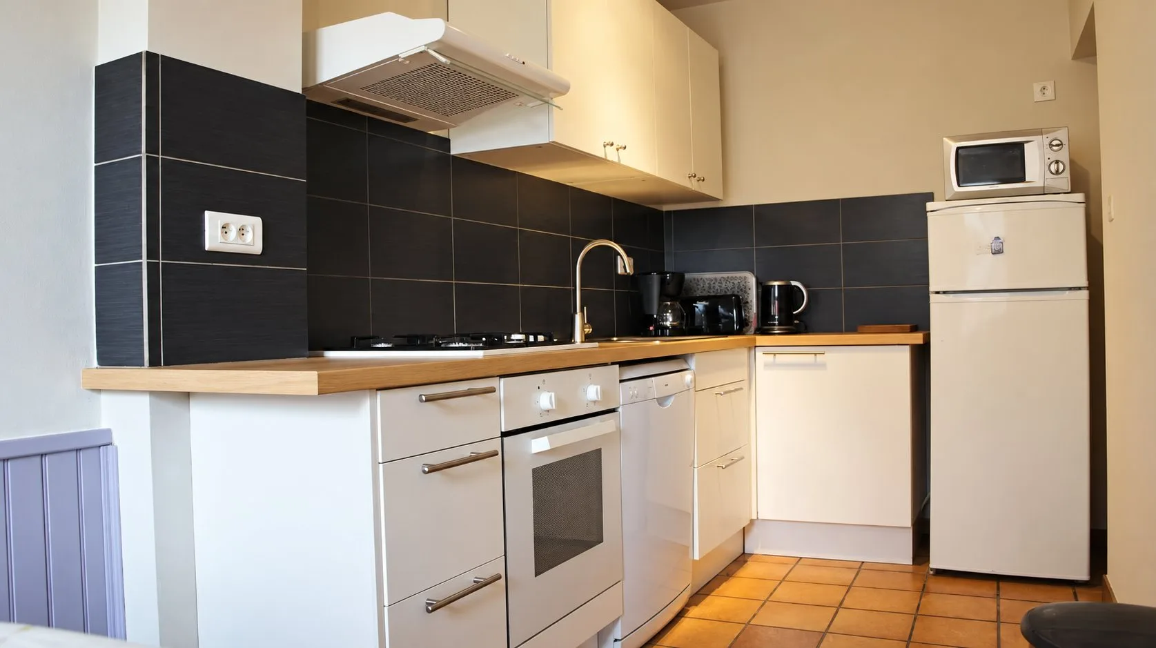 Cuisine en angle avec meubles blancs, plan de travail en bois, crédence anthracite, four, lave-vaisselle, réfrigérateur et micro-ondes.