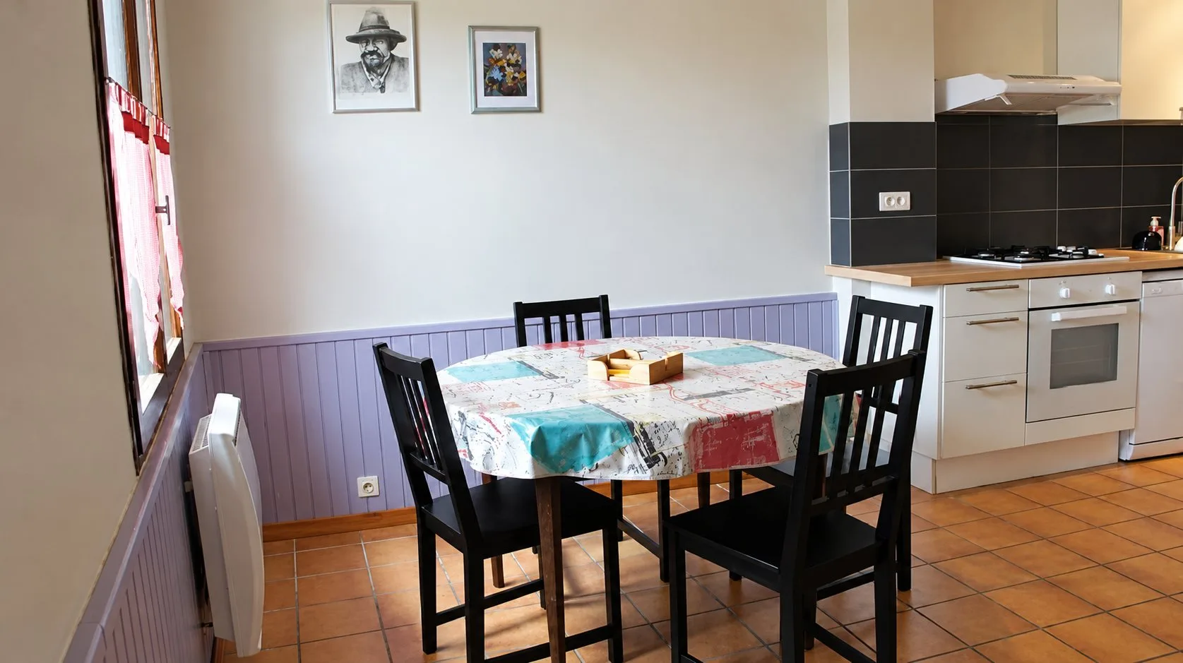 Coin repas d'une cuisine avec une table ronde recouverte d'une nappe colorée, quatre chaises noires, un mur à lambris violet et une cuisine équipée blanche au fond.