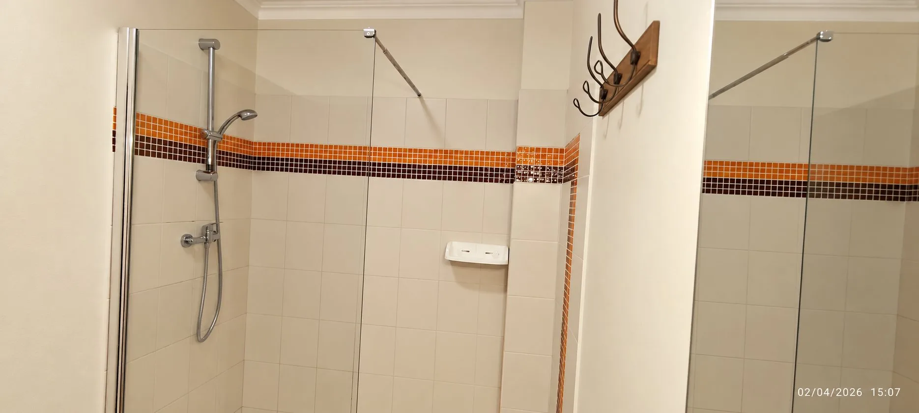 Une salle de bain avec une douche à paroi de verre, une frise de mosaïque orange et brune, une étagère blanche et un portemanteau mural.