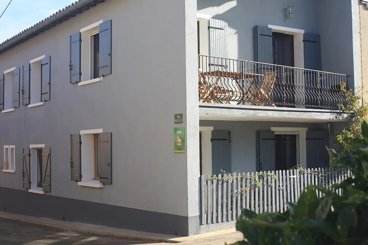 Façade d'une maison grise à volets gris et balcon en fer forgé avec table et chaises en bois, portant une plaque verte indiquant un gîte.