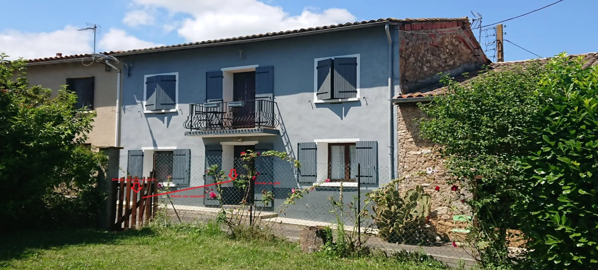 Façade d'une maison de village peinte en bleu-gris, avec volets gris, balcon en fer forgé et jardin au premier plan, des annotations rouges figurant sur l'image.