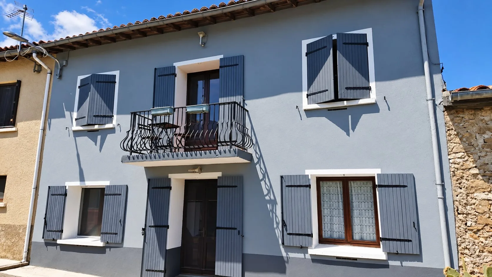 Façade d'une maison de village repeinte en gris, avec volets gris foncé, encadrements blancs et un balcon en fer forgé.