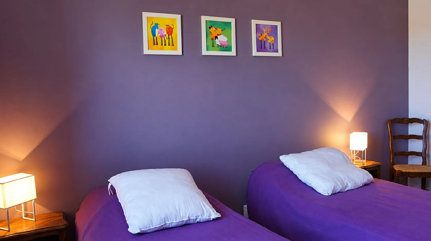 Chambre du gîte Balcon aux murs mauves, avec deux lits simples violets et trois petits tableaux colorés.