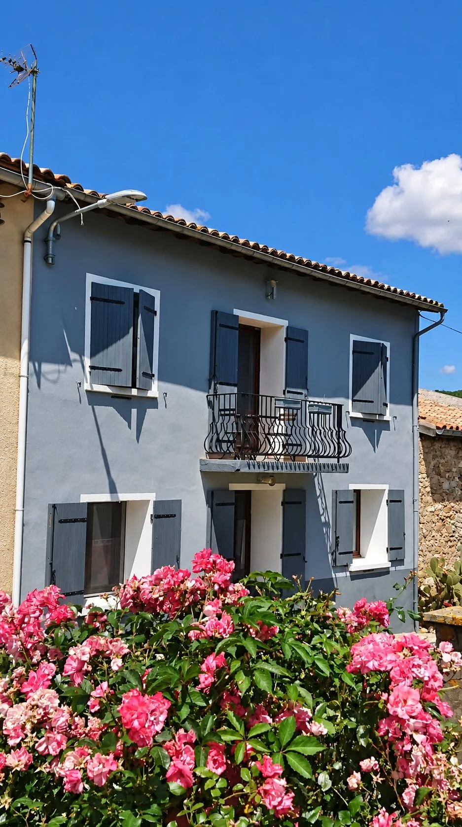 Façade grise du gîte Balcon au-dessus d'un massif de rosiers roses en fleurs.