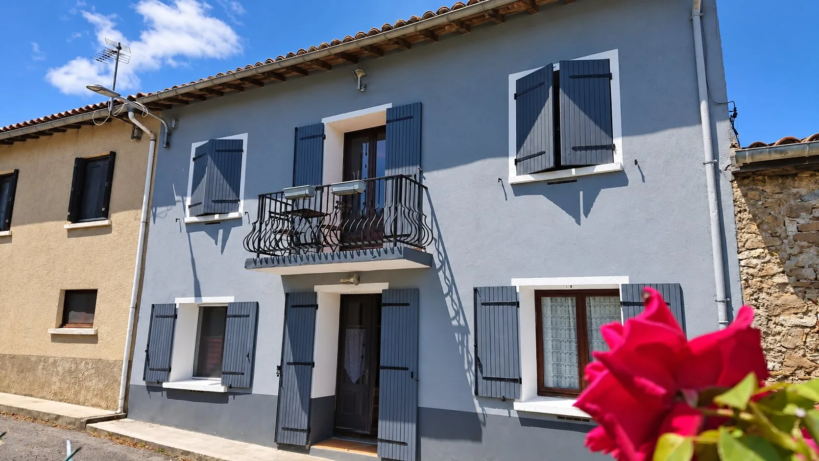 Façade grise aux volets bleu-gris du gîte Balcon, avec son balcon en fer forgé et une rose rouge au premier plan.