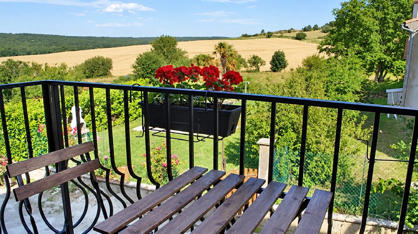 Vue depuis le balcon en fer forgé du gîte Balcon, avec un banc en bois, une jardinière de géraniums et des prairies au loin.