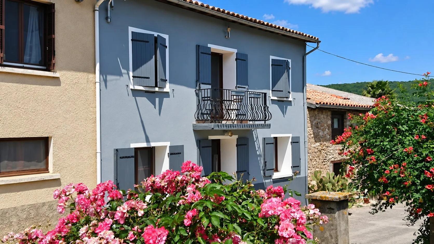 Façade d'une maison bleue avec un balcon en fer forgé et des volets gris, entourée de rosiers roses et d'un arbuste à fleurs rouges sous un ciel bleu.