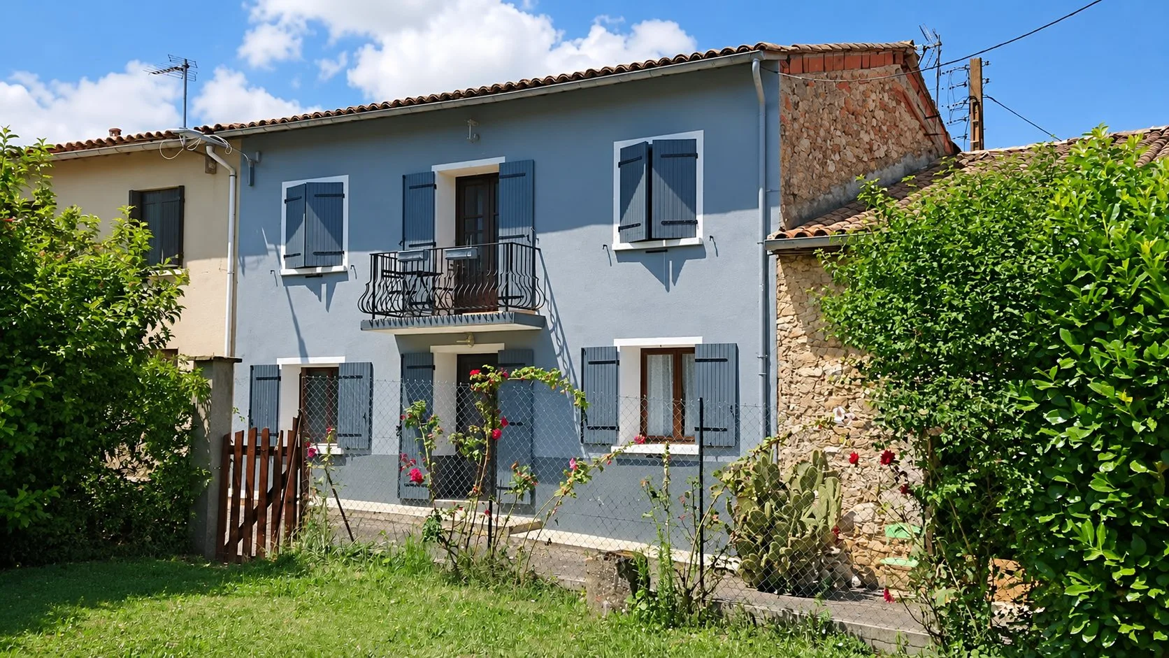 Façade d'une maison de village aux murs gris et aux volets bleu-gris, avec un balcon en fer forgé, entourée de verdure et de rosiers.