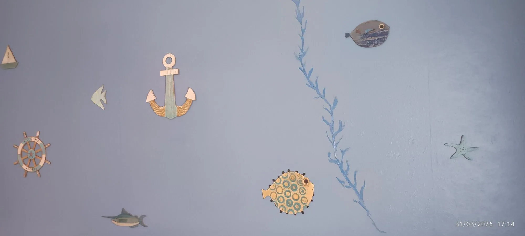 Un mur bleu orné de décorations marines en bois, dont une ancre, un gouvernail, des poissons, une étoile de mer et une algue peinte.