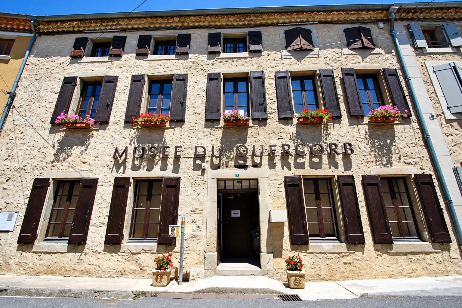 Façade en pierre du musée du Quercorb, avec ses volets bruns et ses jardinières fleuries.