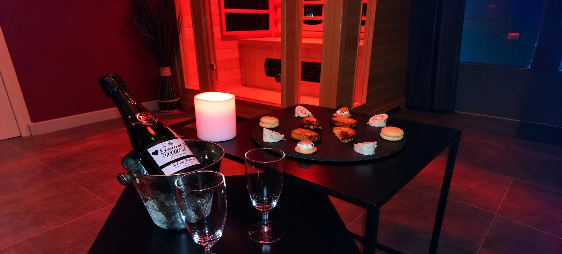 Table de Piccolove avec une bouteille en seau, deux verres, une bougie et un plateau de petits fours, devant le sauna éclairé en rouge.