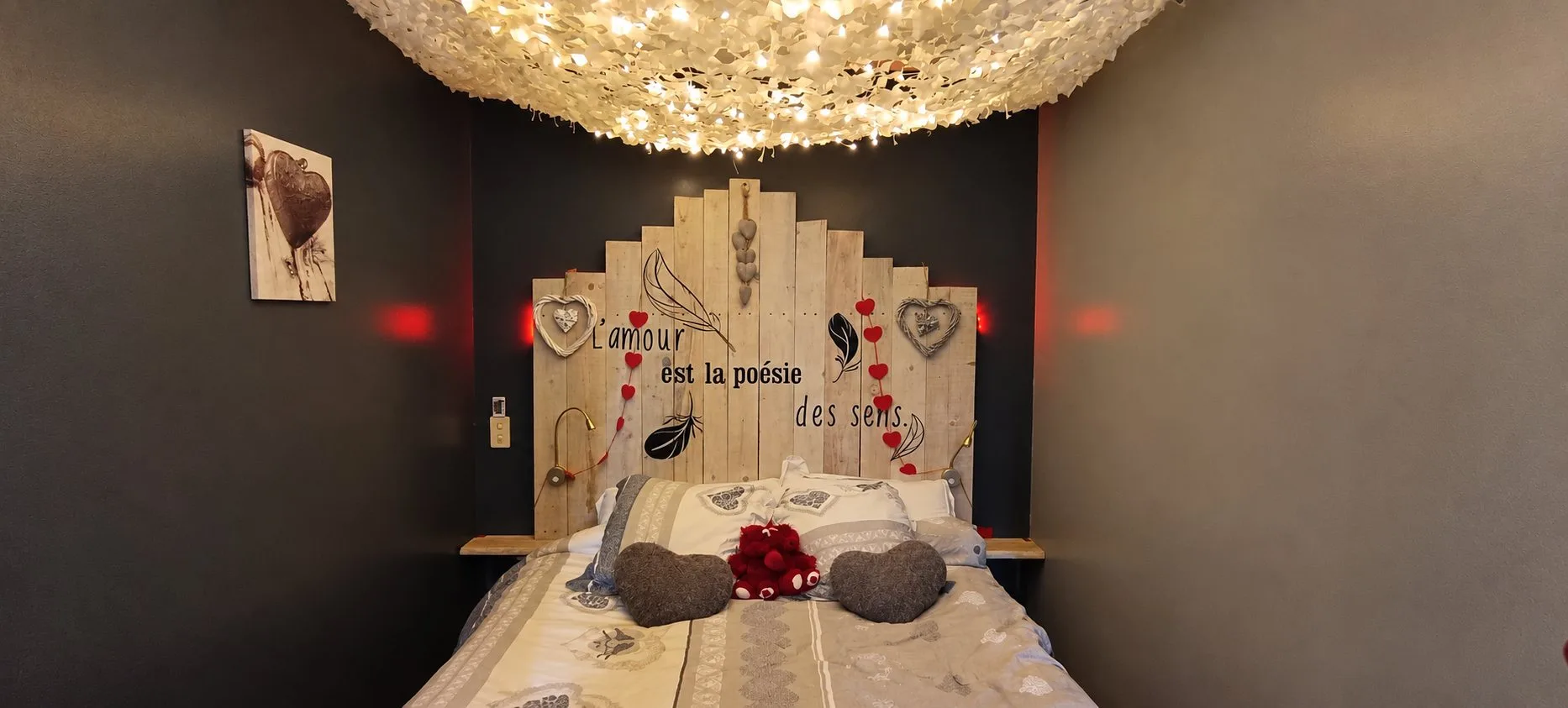 Chambre de Piccolove aux murs sombres, avec une tête de lit en bois de palette ornée de cœurs et un plafonnier lumineux.