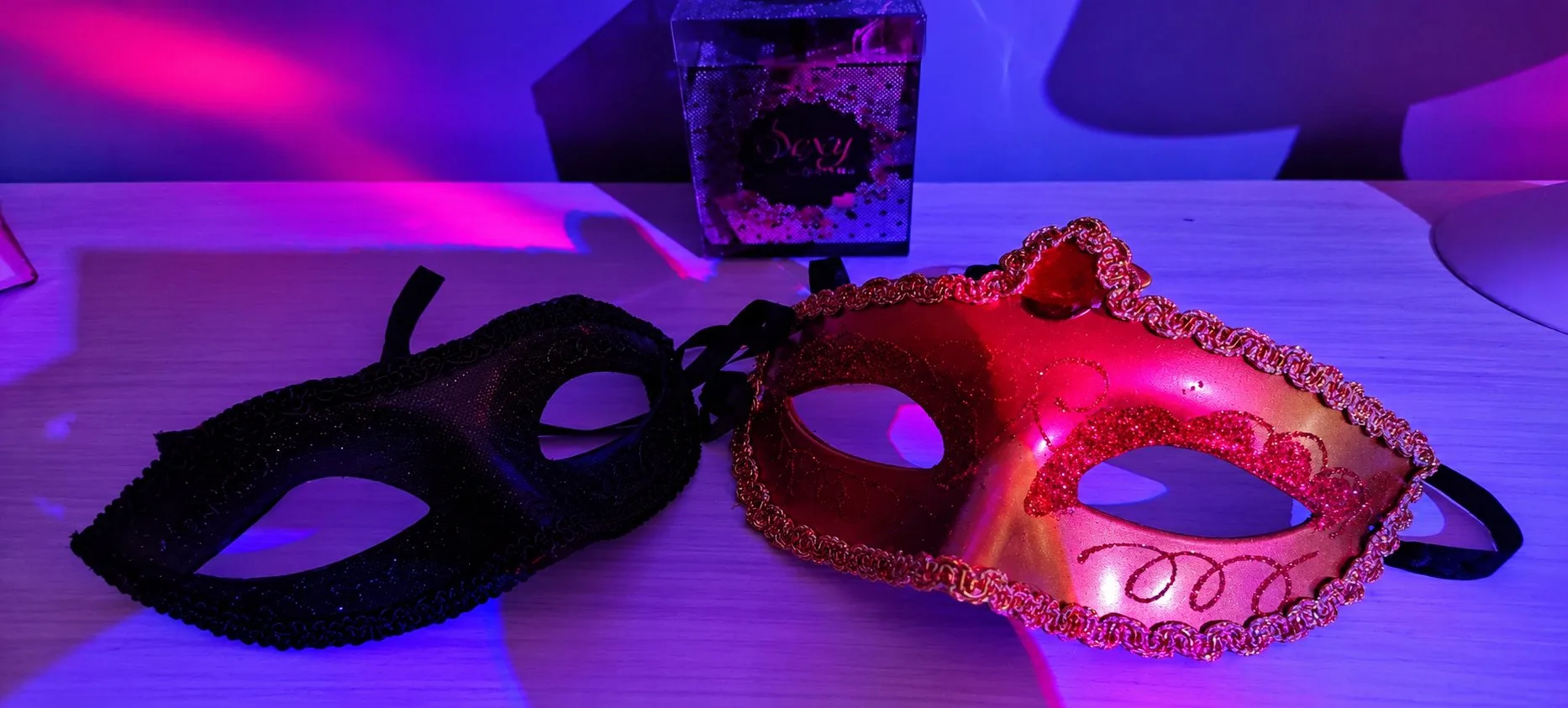 Deux masques vénitiens, l'un noir et l'autre rose pailleté, posés sous une lumière violette.
