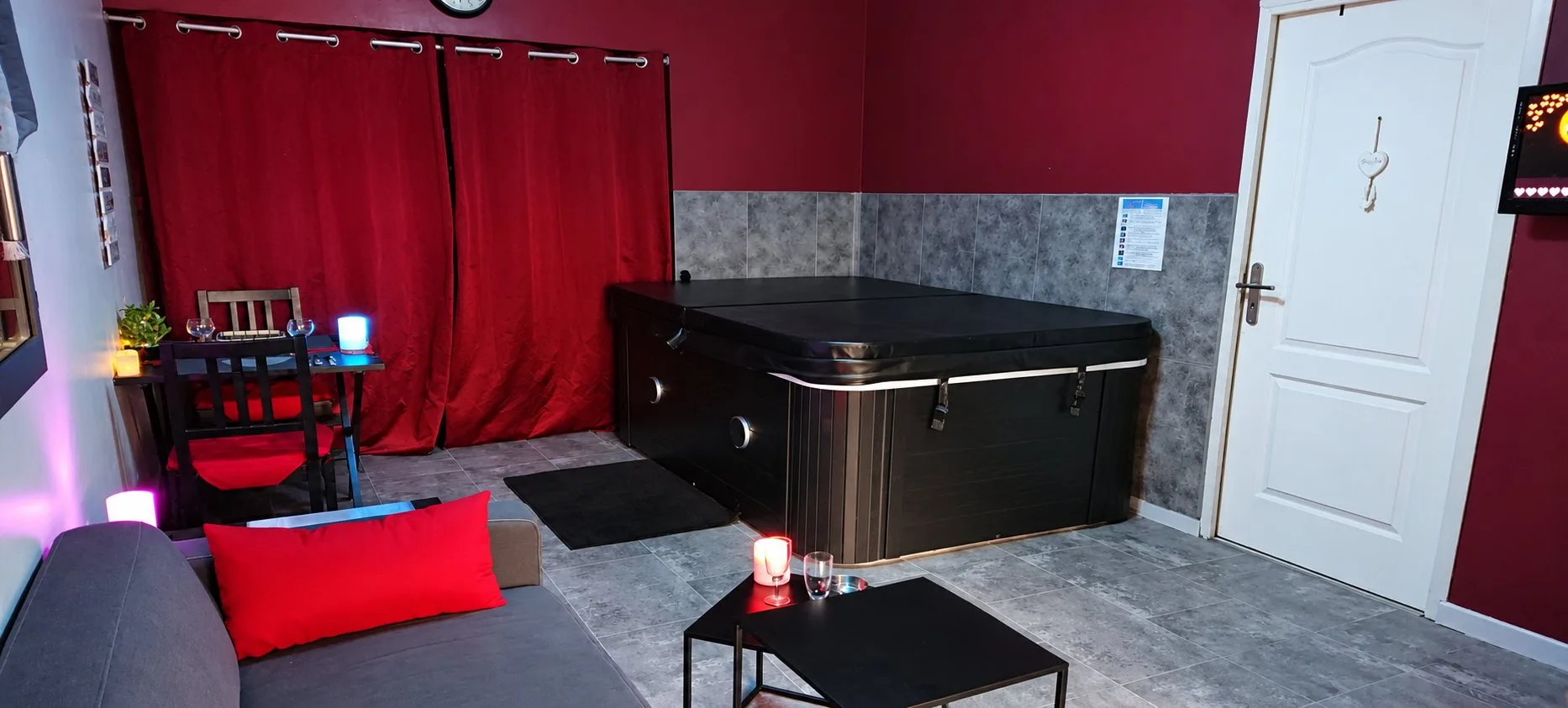 Séjour de Piccolove avec un canapé gris et un coussin rouge, une table de bistrot, des rideaux rouges et le spa sous son couvercle.
