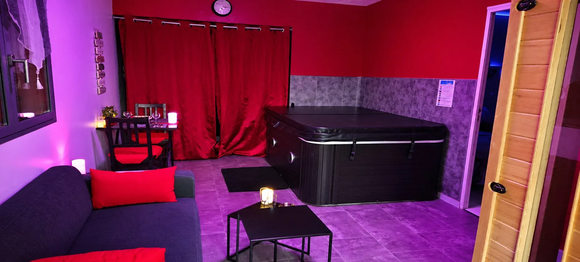 Séjour de Piccolove sous un éclairage violet, avec le canapé, la petite table, le spa et la porte du sauna en bois clair.