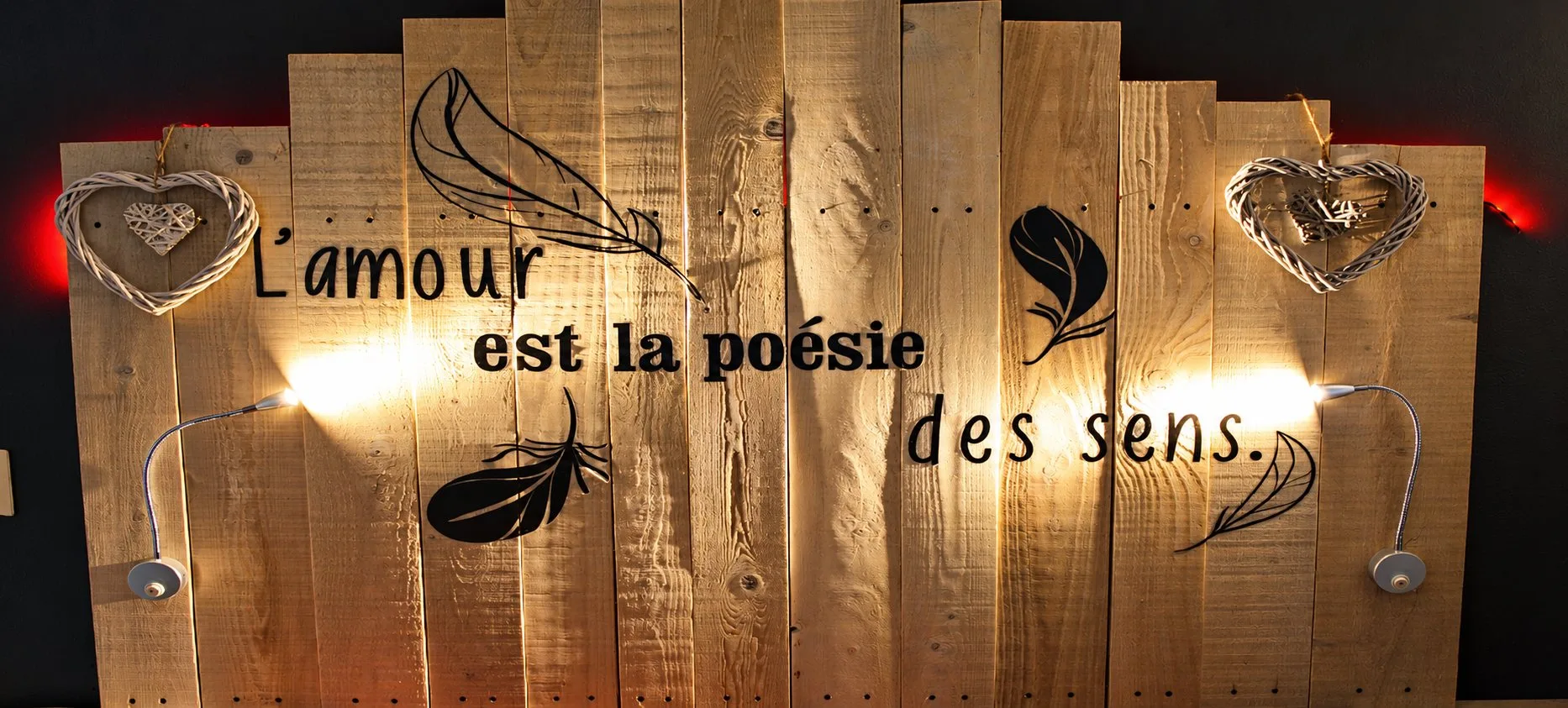 Tête de lit en bois de palette éclairée par deux liseuses, avec des cœurs en osier et des plumes noires.