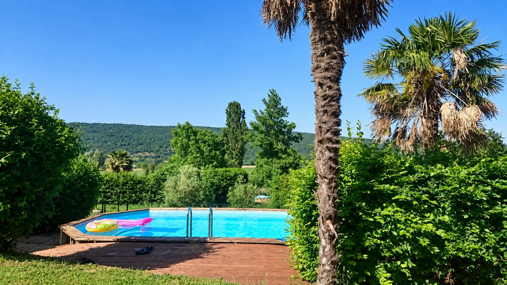 Une piscine en bois au bord d'une terrasse, avec des bouées flottantes, entourée de palmiers, de haies et de collines boisées sous un ciel bleu.