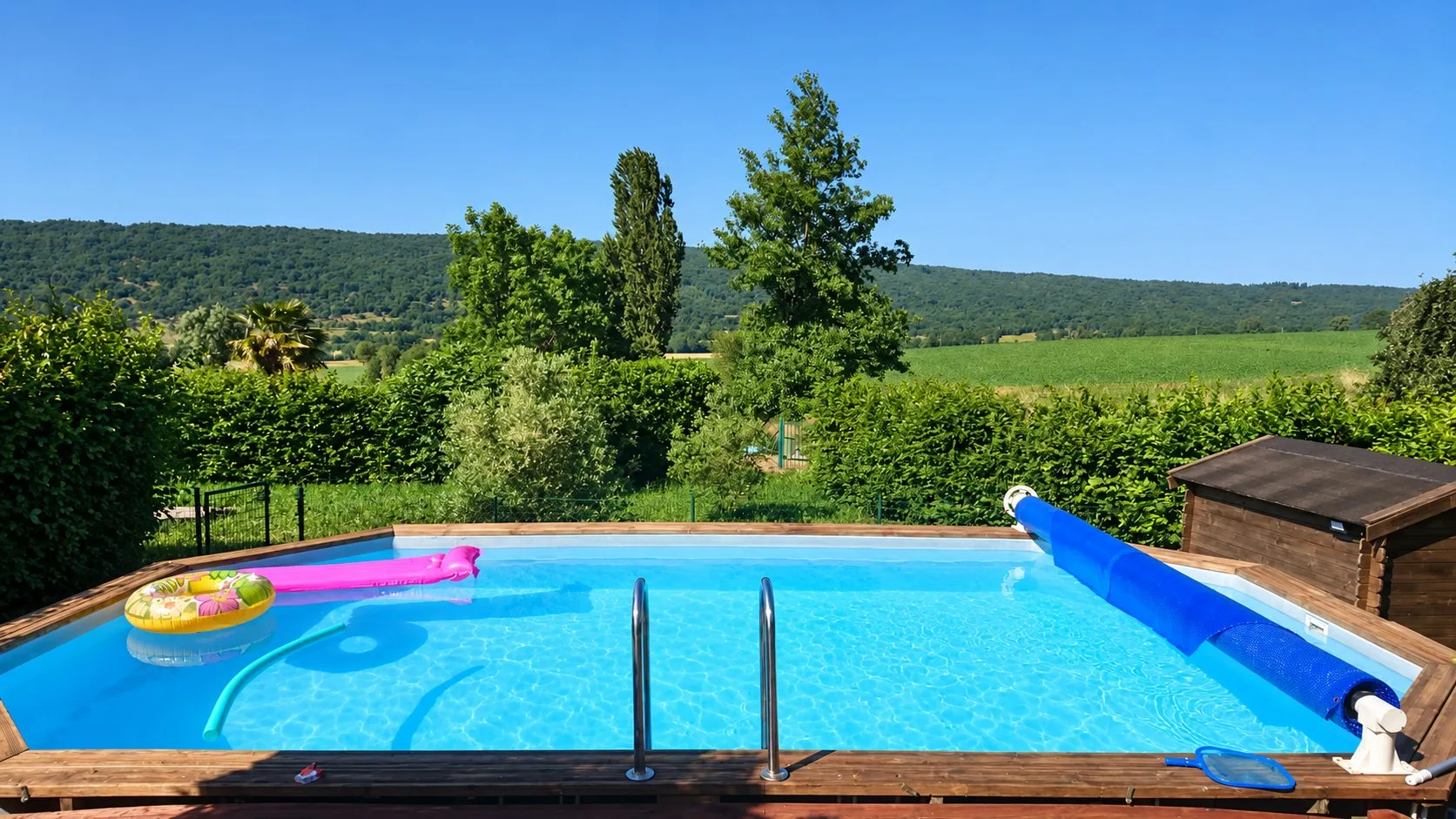 Une piscine hors-sol à margelle en bois, avec bouée, matelas rose et bâche à bulles enroulée, devant une colline boisée et des champs verts.