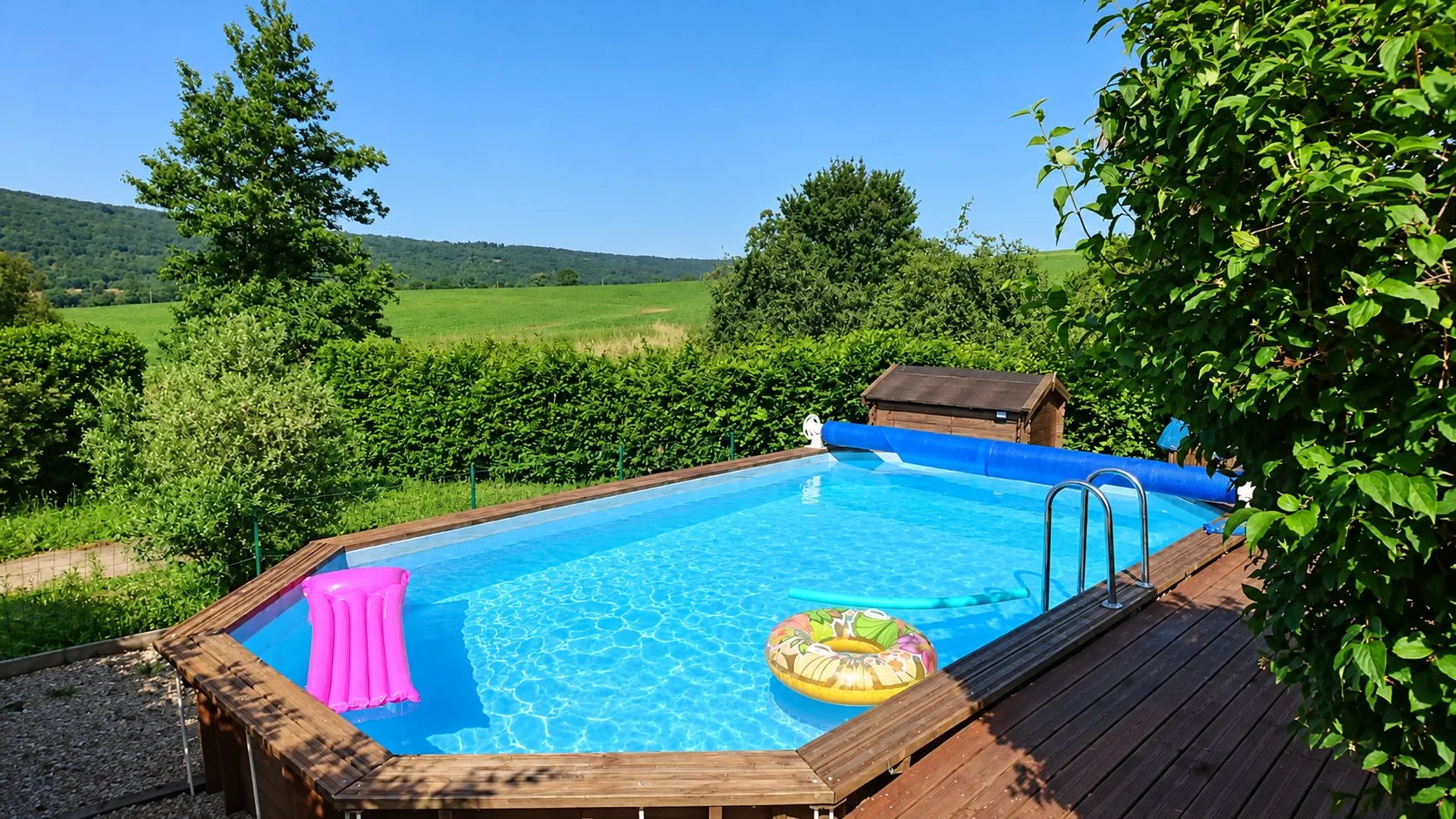 Piscine rectangulaire en bois avec bouée et matelas rose, entourée de haies et d'une terrasse, face à des prairies et collines.