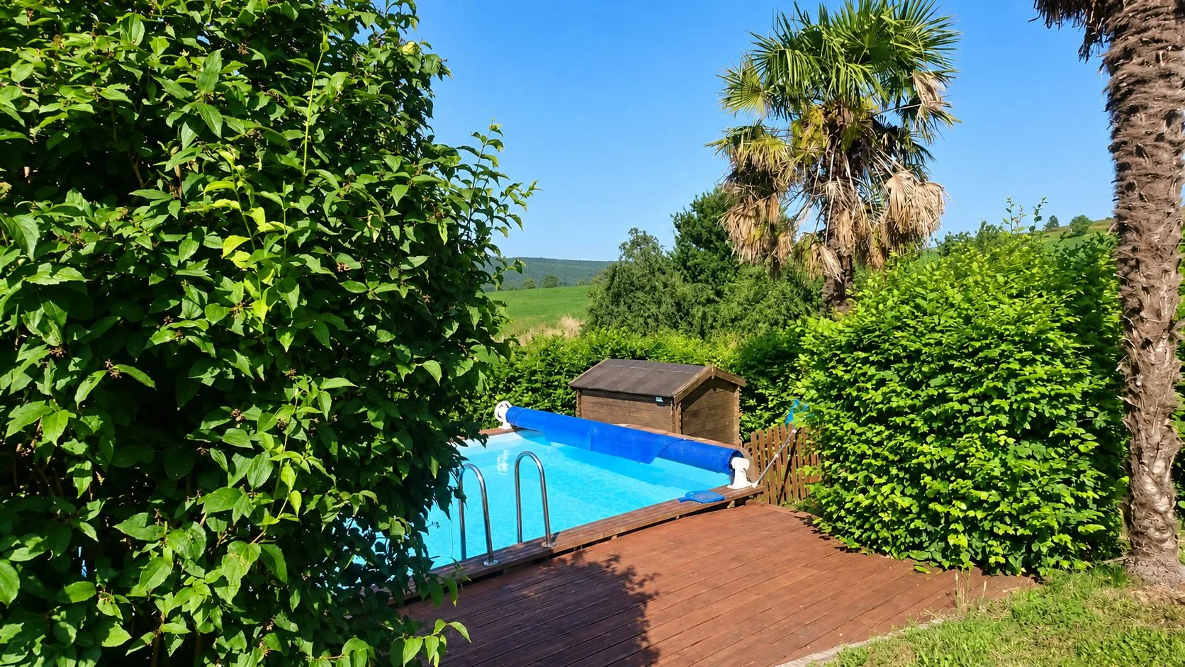 Une piscine rectangulaire avec une bâche à bulles bleue et une terrasse en bois, entourée de haies, d'un palmier et d'un abri de jardin, avec la campagne en arrière-plan.