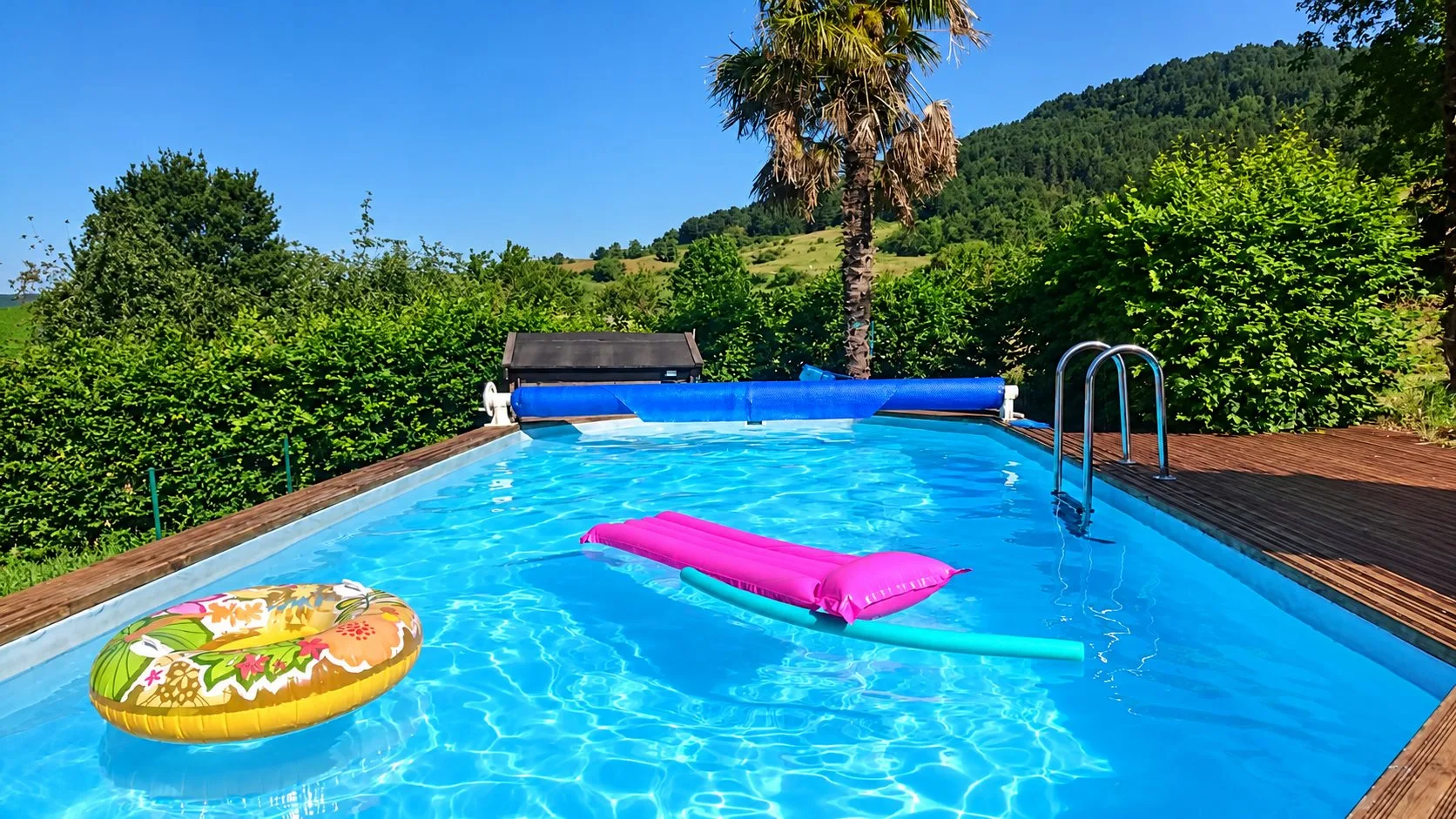 Une piscine rectangulaire à l'eau turquoise, entourée d'une terrasse en bois et de haies, avec une bouée, un matelas rose et une frite flottant à la surface.