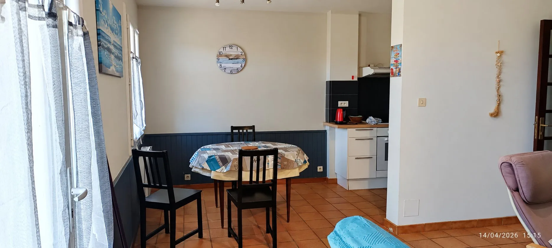 Pièce de vie avec une table ronde et des chaises noires, une horloge murale et une kitchenette blanche et noire au fond, sur un carrelage orangé.