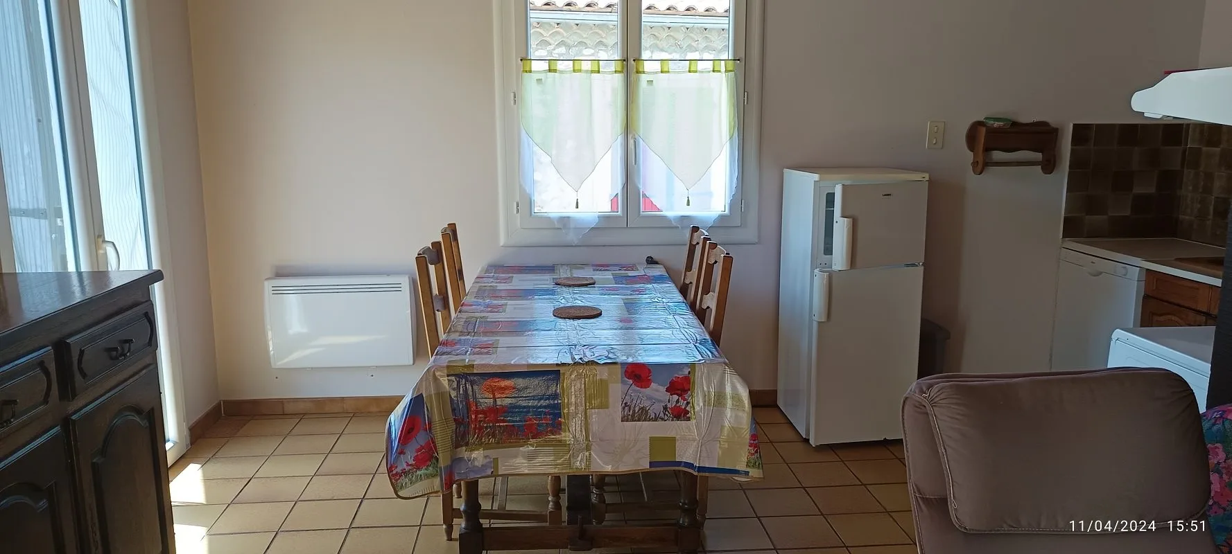 Pièce de vie avec une table recouverte d'une nappe fleurie entourée de chaises en bois, un radiateur mural, un réfrigérateur blanc et une kitchenette.