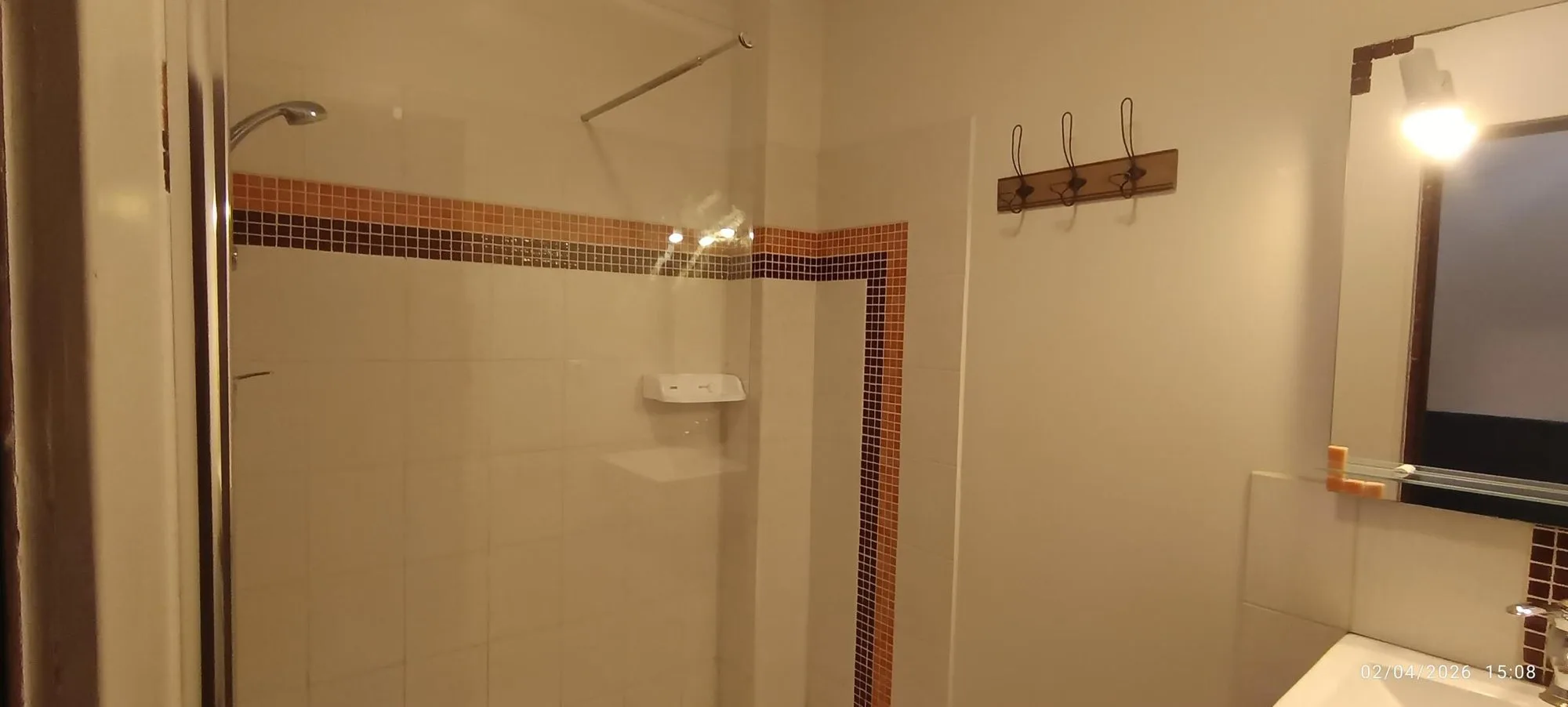 Salle de bain aux murs clairs avec une douche à paroi de verre, une frise de mosaïque orange et brune, un porte-manteau mural et un miroir éclairé au-dessus du lavabo.