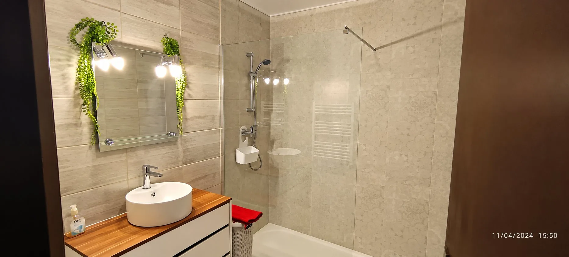 Salle de bain moderne en carrelage beige avec meuble vasque en bois, miroir éclairé et douche à l'italienne avec paroi de verre.