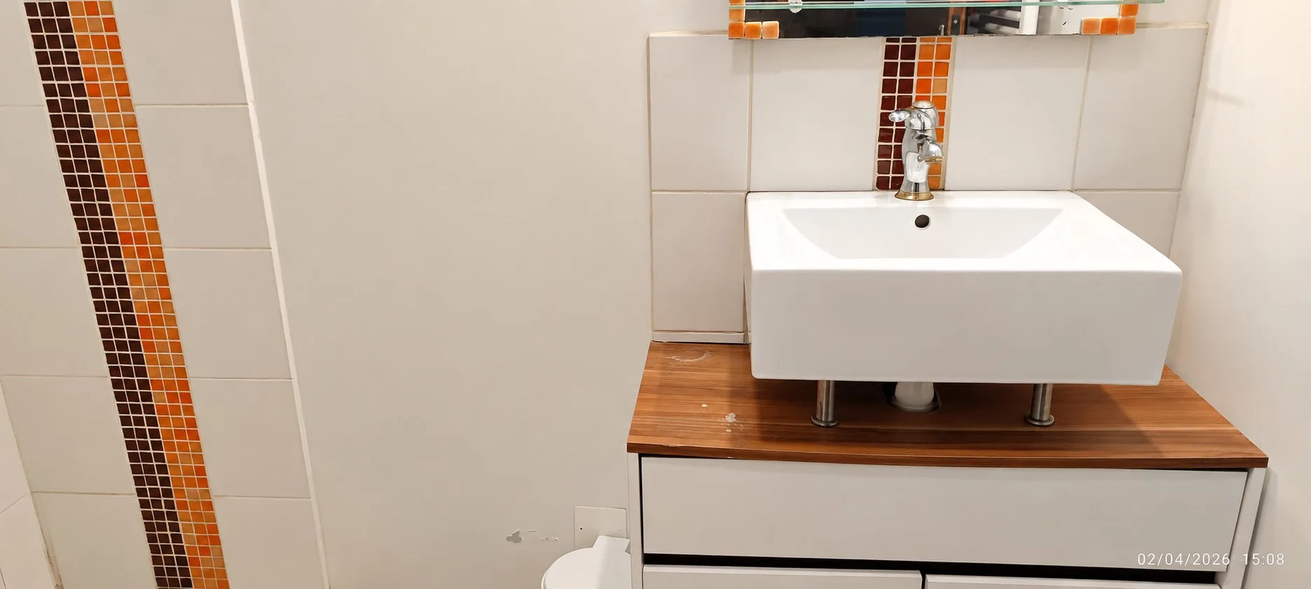 Une vasque blanche carrée posée sur un meuble en bois avec un mitigeur chromé, à côté d'une frise de mosaïque dans les tons orange et marron sur un mur carrelé beige.