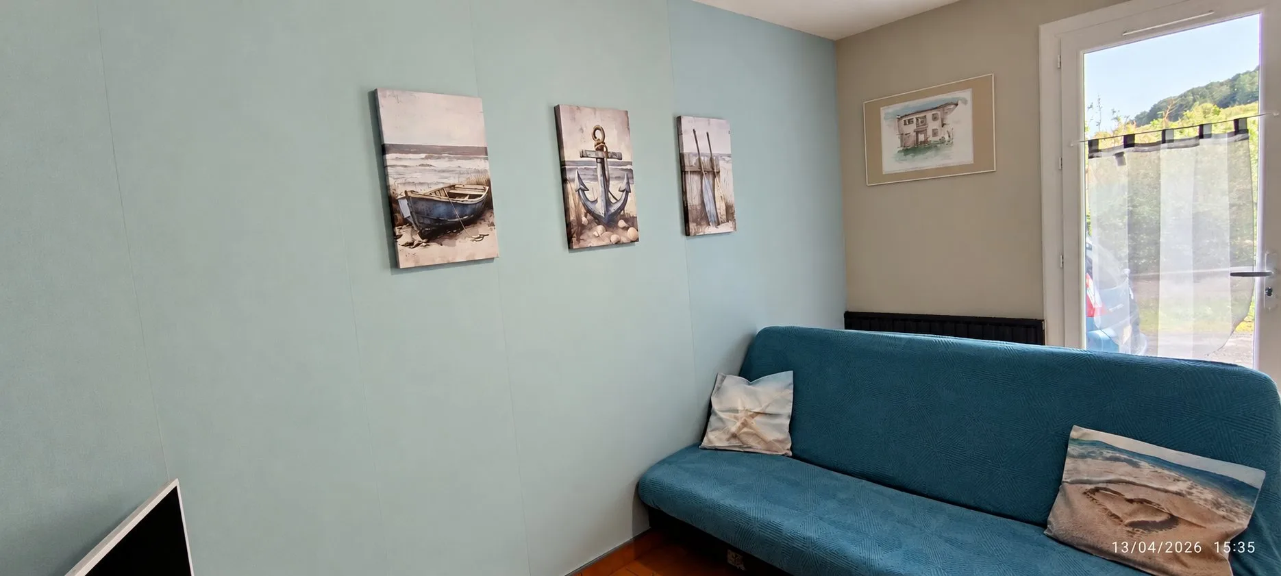 Pièce aux murs vert-bleu décorée de trois tableaux marins, avec un canapé-lit bleu et une porte-fenêtre donnant sur l'extérieur.